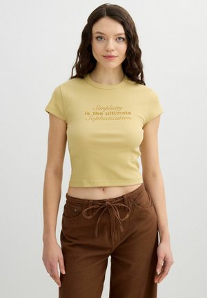 Mujer con cabello largo y rizado lleva una camiseta amarilla corta que dice "La simplicidad es la máxima sofisticación" y pantalones marrones con la cintura atada.