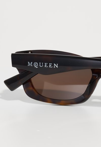 Türi kinnitusprillid pruuni objektiiviga, millel on stiilne must raam ja hõbedane "McQueen" logo ülemisel käeosal.