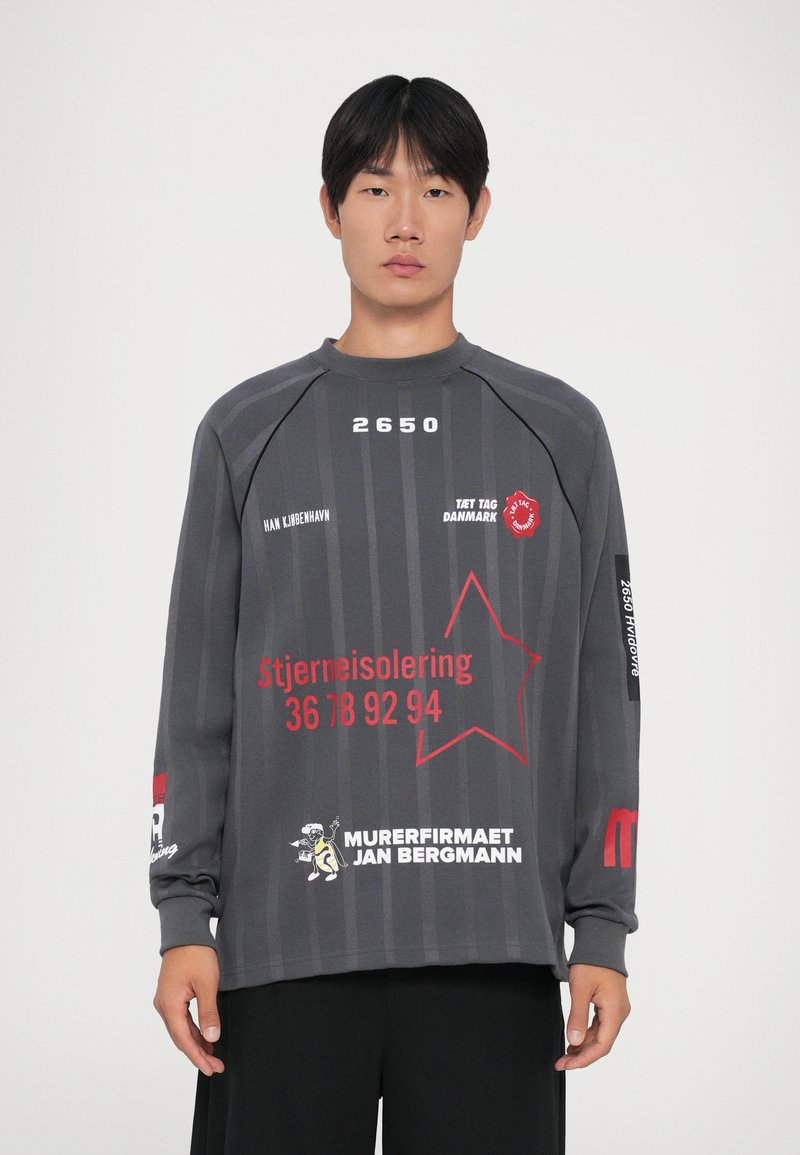 Jovem vestido com uma camisa de manga longa cinza-escura, com texto e gráficos em dinamarquês vermelho e branco, de pé contra um fundo claro e simples.