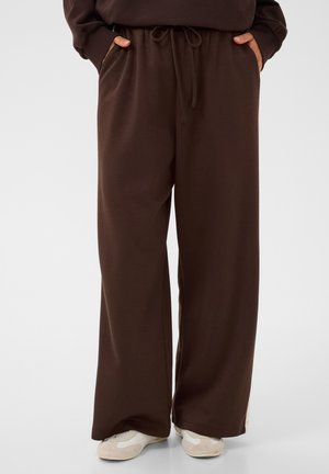 Personne portant un pantalon ample marron foncé avec un cordon à la taille et des baskets blanches, mains dans les poches, debout sur un fond clair uni.