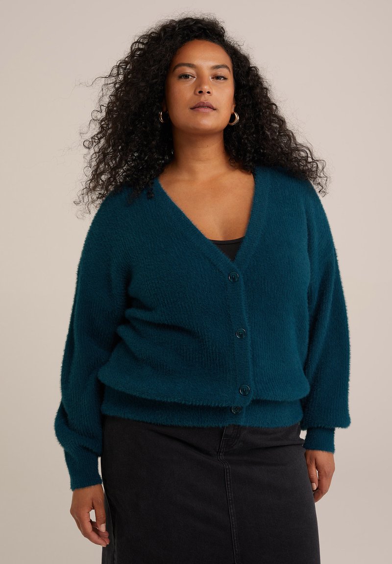 Teal fluffy cardigan med V-ringning, fem knappar och långa ärmar. Buren över en svart topp, tillsammans med en svart kjol. Mjuk textur.