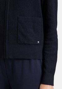 Marineblaues Zip-Up-Jackett aus strukturiertem Stoff, mit gerippten Bündchen und einer praktischen Vordertasche mit metallischem Akzent.