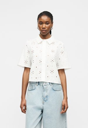 Femme aux cheveux tressés portant un chemisier blanc à manches courtes ajouré et un jean taille haute bleu clair, debout devant un fond uni.