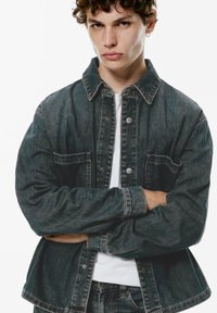 Donkerdenim jack met een knoopsluiting aan de voorkant, spitse kraag en twee borstzakken. De stof heeft een licht versleten textuur en een relaxte pasvorm.