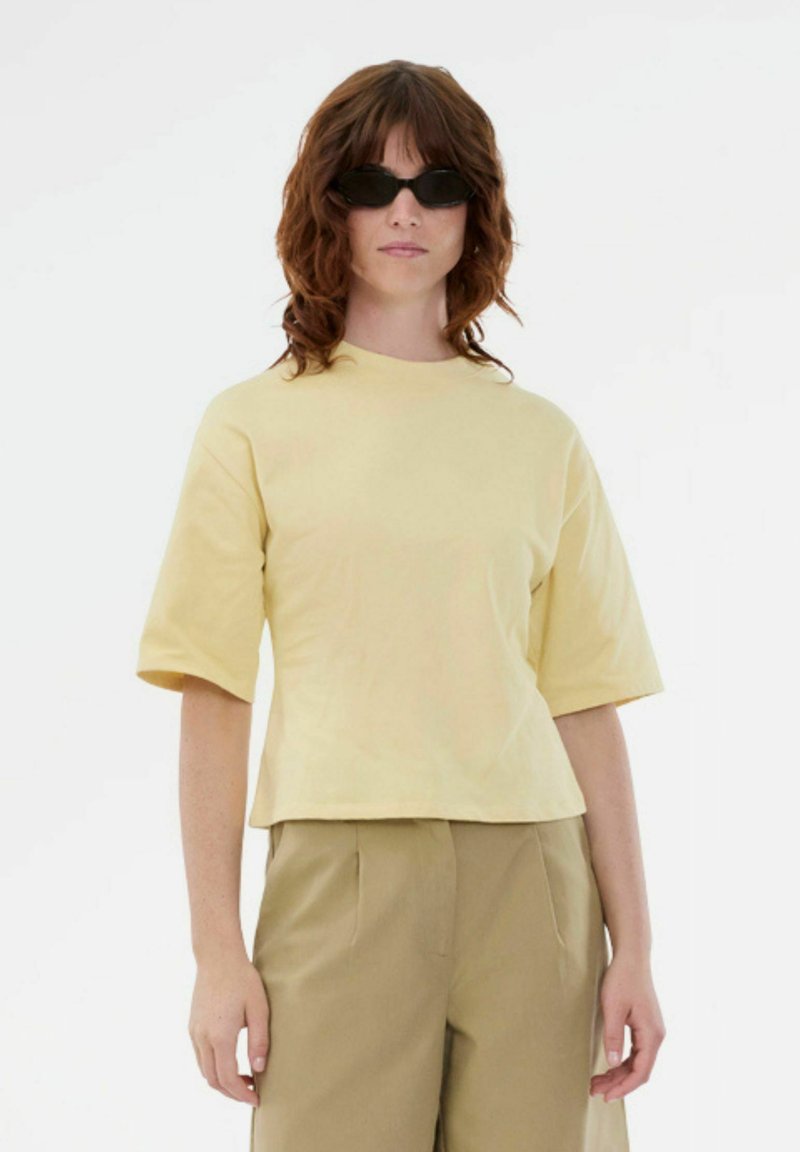 Femeie cu păr castaniu ondulat, lungime până la umeri, purtând ochelari de soare negri, o cămașă scurtă cu mâneci galben deschis și pantaloni bej, stând pe un fundal alb.