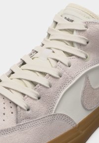 Gros plan d'une chaussure en daim beige avec des lacets blancs, le logo Nike swoosh sur le côté et une semelle en caoutchouc gomme.