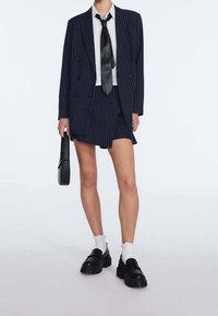 Laivastonsininen pinstripe-bleiseri kaksirivisellä napituksella, yhdistettynä samaa kangasta olevaan lyhyeen hameeseen, valkoiseen paitaan ja mustaan kapeaan solmioon. Paksut mustat loafers-kengät ja sukat.