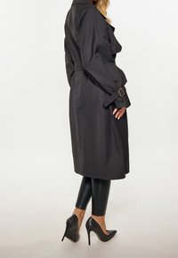Trench-coat long noir avec une ceinture à la taille, des détails à boutons et des poignets retroussés, associé à des leggings noirs brillants et des escarpins à bouts pointus ornés de clous.