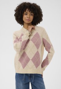 Ženska s kodrastimi lasmi, oblečena v be�ž in roza argyle pulover ter modre kavbojke, pozira z eno roko ob bradi in drugo na boku.