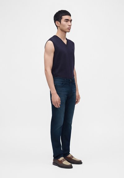 Jeune homme debout, tourné vers la droite, portant un haut sans manches bleu marine foncé, un jean bleu foncé et des chaussures en cuir tressé marron.