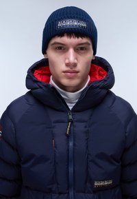 Giacca puffer blu navy con interno rosso, tasche con zip e accenti del logo. Il modello indossa un berretto blu navy a trama su uno sfondo neutro.