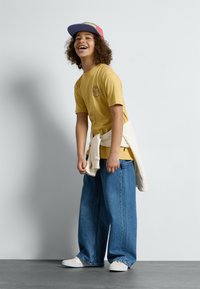 Gelbes kurzärmliges T-Shirt mit einem runden Logo, lässige blaue Denim-Jeans, weiße Sneaker und eine mehrfarbige Kappe. Pullover um die Taille gebunden.