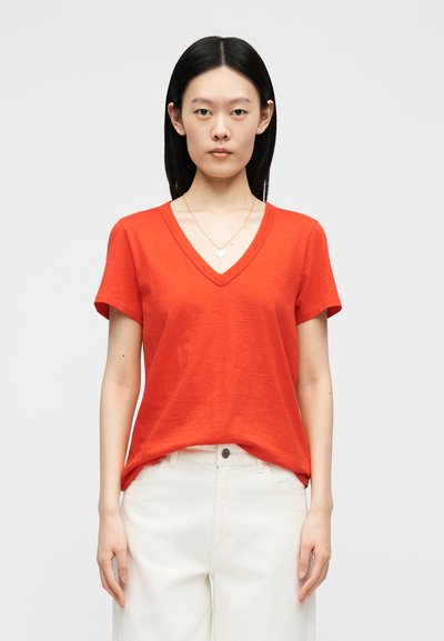 Νέο άτομο με ίσια μαύρα μαλλιά που φοράει ένα φωτεινό κόκκινο κοντομάνικο μπλουζάκι με V λαιμόκοψη και άσπρο παντελόνι, στέκεται μπροστά σε ένα απλό φωτεινό φόντο.