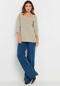 Helles beiges Blusenshirt mit einem runden Ausschnitt, Knopfleiste, gefältelten Details und drei Viertelärmeln, kombiniert mit blauen weit geschnittenen Jeans.
