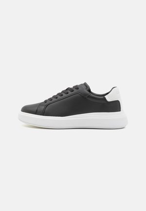 Baskets basses noires avec un patch blanc au talon et une épaisse semelle blanche, avec des lacets noirs et des détails de couture minimalistes.