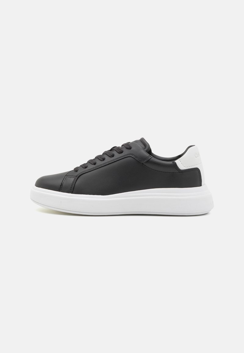 Baskets basses noires avec un patch blanc au talon et une épaisse semelle blanche, avec des lacets noirs et des détails de couture minimalistes.