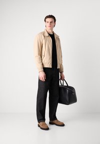Chaqueta bomber beige, camiseta negra de cuello redondo, pantalones vaqueros oscuros, mocasines marrones, sosteniendo un bolso de cuero negro con dos asas.