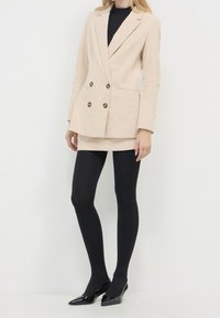 Beżowa dwurzędowa marynarka ze sztruksu o teksturowanej powierzchni, zestawiona z beżową mini spódnicą, czarnymi rajstopami i czarnymi szpilkami z ostrym noskiem.