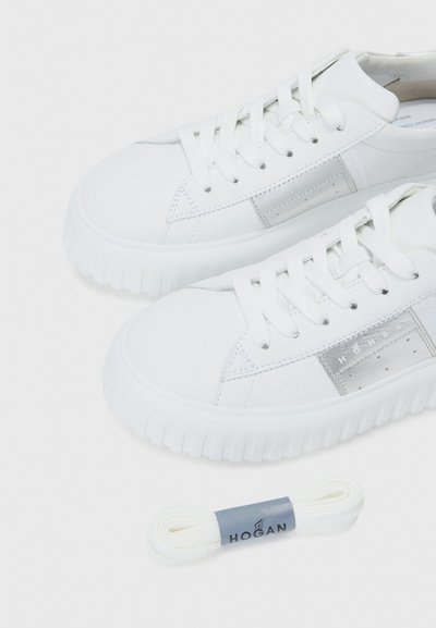 Baskets blanches à tige basse avec des semelles épaisses et des lacets blancs, présentant des empiècements latéraux argentés et une paire supplémentaire de lacets blancs de marque en dessous.
