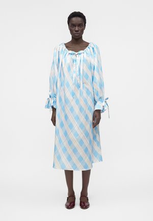 Kobieta ubrana w luźną, midi sukienkę z długim rękawem w niebiesko-białą, ukośną kratę, z kokardkami przy dekolcie i mankietach, połączoną z ciemnoczerwonymi butami.