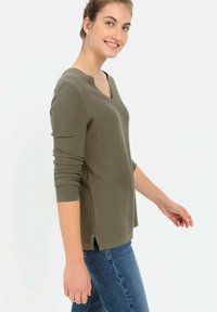 Oliwkowy sweter z dekoltem w serek i długimi rękawami, o fakturze rib, z rozcięciami po bokach u dołu, noszony na niebieskie dżinsy. Luźny krój.