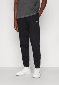 Pantalones deportivos atléticos negros hechos de tela suave, con un ajuste ceñido y cintura elástica. Un pequeño logo de Reebok está impreso en el muslo izquierdo.