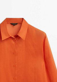 Chemise orange vif à manches longues avec col pointu et boutons sur le devant, en tissu léger texturé.