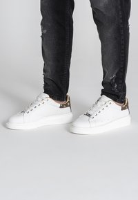 Weiße Sneakers mit flacher Sohle und beigen gemusterten Akzenten an der Ferse, ausgestattet mit sechs Ösen und strukturierten Textilelementen.