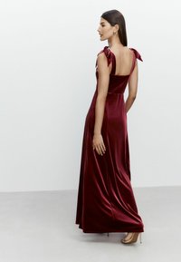 Rochie maxi din catifea burgundy, cu bretele legate în fundiță, spate adânc și fustă fluidă. Textură netedă și siluetă ajustată.