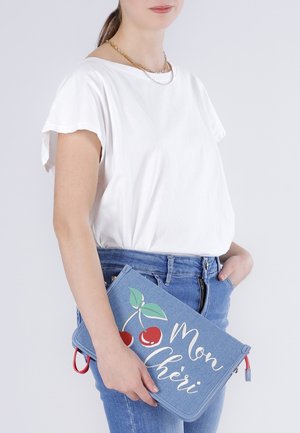 Vrouw in wit shirt en blauwe spijkerbroek die een blauwe clutch vasthoudt met een kersontwerp en de tekst "Mon Chéri".