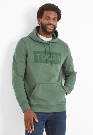 Green hoodie with front pocket, drawstring hood, and embossed "TOG24" logo. Fabric shows a smooth texture and casual fit. Worn over a white shirt.