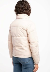 Chaqueta acolchada en beige claro con cuello alto, textura acolchada, puños elásticos y bolsillos laterales, mostrada desde la parte posterior.