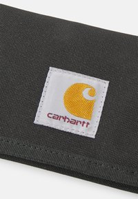 Black fabric wallet with textured surface; features a white square patch with a yellow logo and "carhartt" text in red.