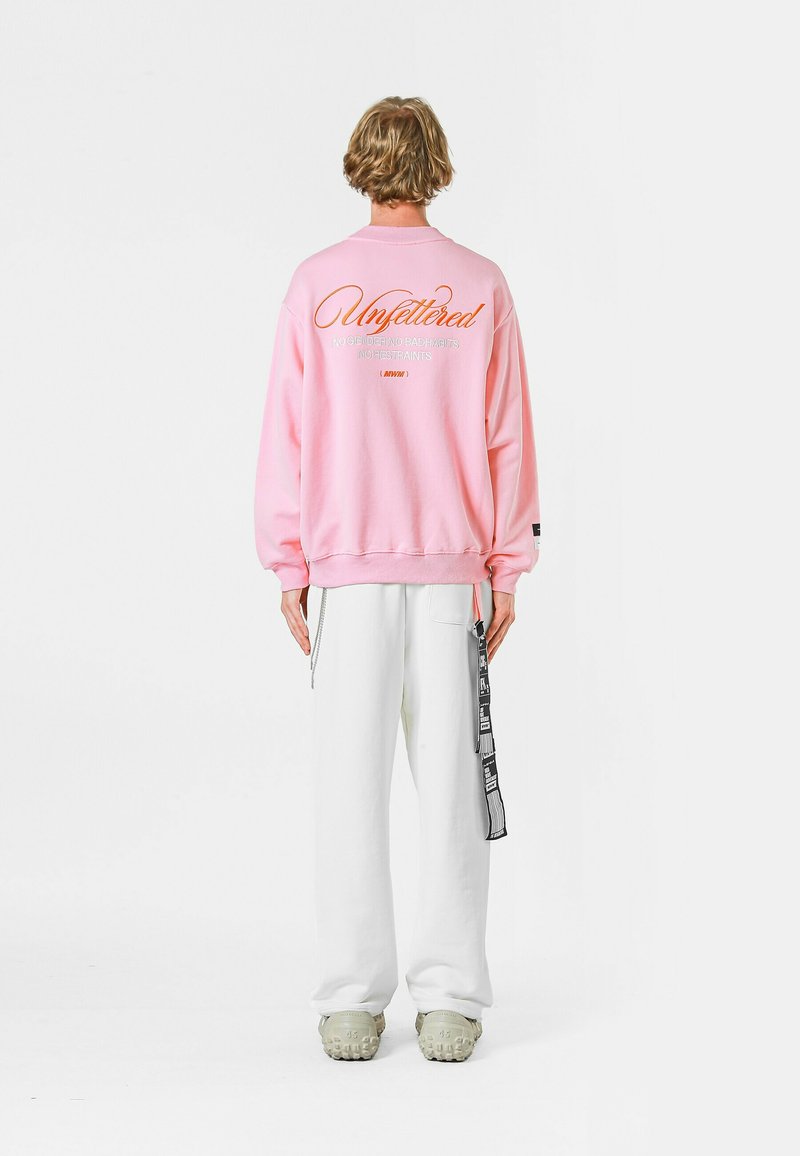 Sudadera rosa con el texto "Sin restricciones" en la parte posterior, combinada con pantalones anchos blancos y calzado beige con suelas texturizadas.