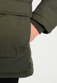 Ciemnozielona kurtka puchowa z boczną kieszenią, o gładkiej fakturze i przeszyciach. Zawiera metkę z logo u dołu.