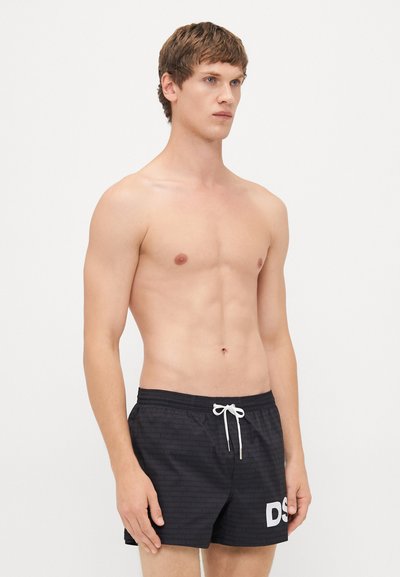 Junger Mann mit kurzen braunen Haaren, der schwarze Badeshorts mit weißen "DS"-Buchstaben am Bein trägt, steht vor einem schlichten hellen Hintergrund.