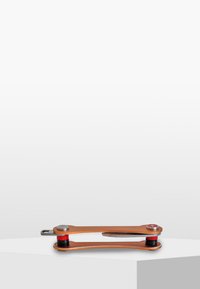 Multiverktyg med en kopparfärgad kropp, röda gummidetaljer och silverfärgade fästen. Har en platt kant och rundade ändar för grepp. Slät yta.