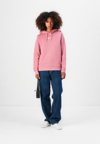 Sweat à capuche rose avec logo sur le devant, associé à un jean bleu ample et des baskets blanches. Modèle tenant un petit sac noir.