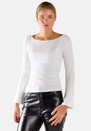 Donna che indossa un top bianco aderente a maniche lunghe e pantaloni in pelle neri, in piedi davanti a uno sfondo chiaro semplice.