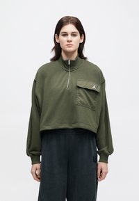 Άτομο που φοράει μια ελαιοπράσινη κοντομάνικη μπλούζα με φερμουάρ και καπιτονέ τσέπη, καθώς και μαύρο φαρδύ παντελόνι, μπροστά από λευκό φόντο.