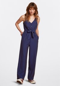 Tutina a righe blu navy con scollatura a V, bottoni frontali, vita con cintura e gambe larghe. Il tessuto ha una consistenza liscia.