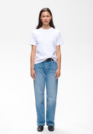 Jeune femme debout, face à l’avant, portant un t-shirt blanc uni, un jean bleu clair avec une ceinture noire et des chaussures noires sur fond blanc.