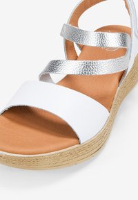 Sandalia blanca con una amplia tira en los dedos y un diseño de tira cruzada en color plata metálico. Cuenta con una plantilla acolchada en tono marrón y una suela texturizada de tejido.