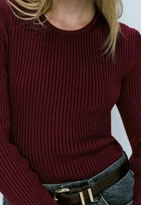 Burgundowa sweterka z prążkowanej dzianiny z długimi rękawami, okrągłym dekoltem i dopasowanym krojem, w zestawieniu z czarnym paskiem i niebieskimi dżinsami.