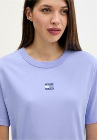 Fialově světle fialové bavlněné tričko s kulatým výstřihem a krátkými rukávy, s modro-bílou nášivkou s logem "TOMMY JEANS" na hrudi.