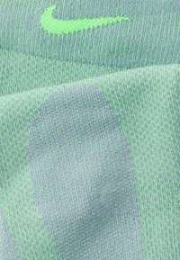 Πράσινο λογότυπο Nike σε υφασμάτινη επιφάνεια ανοιχτού μοβ με υφή, που παρουσιάζει λεπτές, διαπλεκόμενες κλωστές και ποικίλες αποχρώσεις.