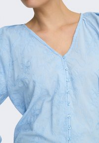 Blouse bleu clair à boutons avec broderie florale portée par une personne, montrant le décolleté et le haut du torse sur un fond uni.