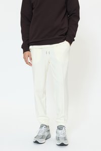 Bela trenirka z ravnim sprednjim delom, elastičnim pasom in zavezovalnim trakom. Kombinirana s temno rjavim puloverjem in sivimi supergami.