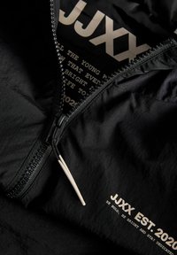 Melna jaka ar daļēji atvērtu rāvējslēdzēju, kurā redzams balts teksts “JJXX EST. 2020” un sauklis “Esi drosmīgs, esi gaišs un paliec uzticīgs.”