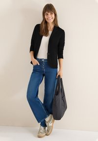 Schwarze Blazer mit zwei Knöpfen über einem weißen Oberteil, kombiniert mit blauen Jeans und hellolivfarbenen Sneakers. Hält eine schwarze Schultertasche.